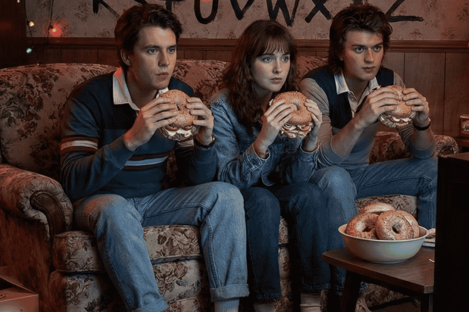 Stranger Things crew sitting on a retro couch in an 80s-style living room eating bagel sandwiches.