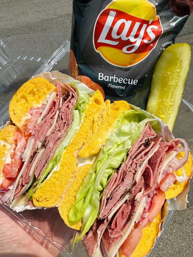 Fresh pastrami deli sandwich on a colored bagel served with pickle and barbecue chips at Fast Bagel Milltown.