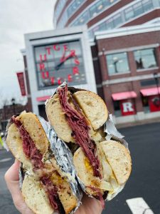 A hand holding a stacked meat, egg, and cheese bagel sandwich wrapped in foil in front of the Rutgers University clock tower in New Brunswick.