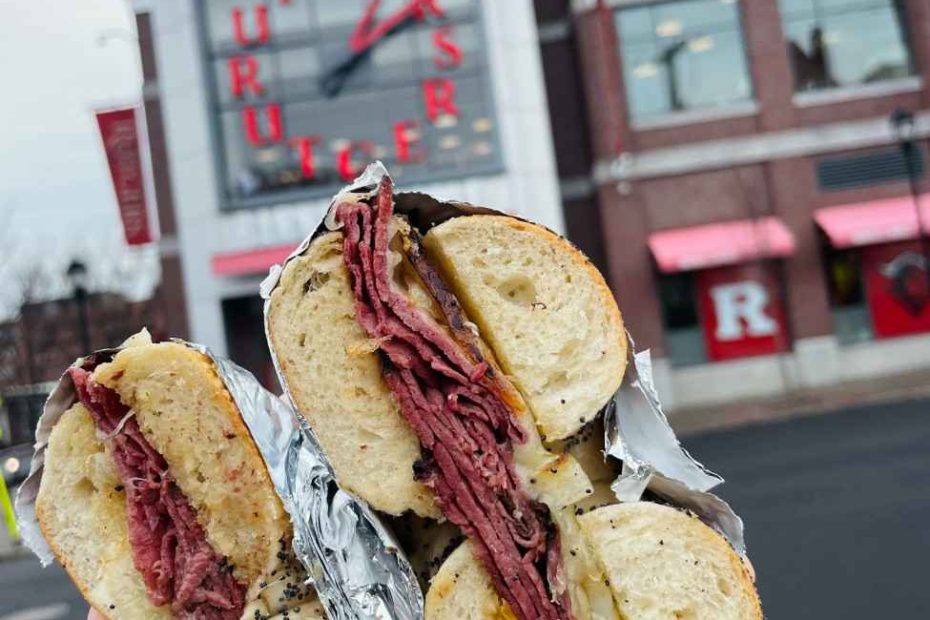 A hand holding a stacked meat, egg, and cheese bagel sandwich wrapped in foil in front of the Rutgers University clock tower in New Brunswick.