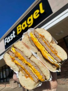 A hand holding a thick sausage, fresh egg, and cheese breakfast sandwich cut in half in front of the Fast Bagel storefront sign.