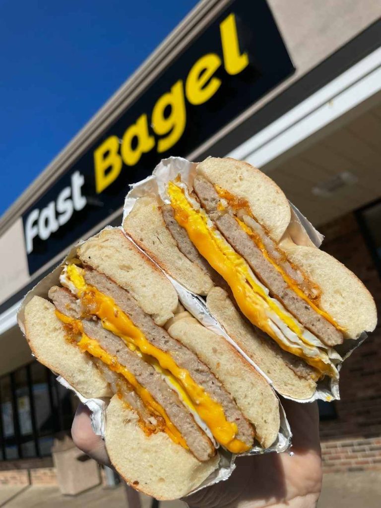 A hand holding a thick sausage, fresh egg, and cheese breakfast sandwich cut in half in front of the Fast Bagel storefront sign.