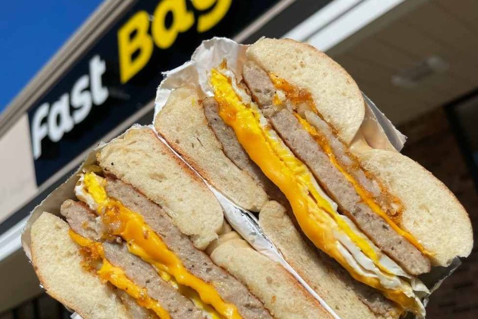 A hand holding a thick sausage, fresh egg, and cheese breakfast sandwich cut in half in front of the Fast Bagel storefront sign.
