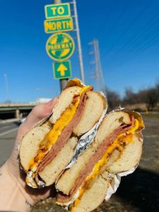 A fresh Pork Roll, egg, and cheese NJ breakfast sandwich held in front of a Garden State Parkway sign.
